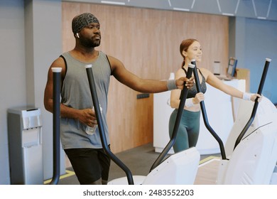 Biracial gym residents running on elliptical machine, pretty woman enjoying process, and young African American man listening to music - Powered by Shutterstock