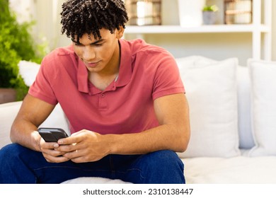 Biracial handsome young man texting over mobile phone while sitting on sofa in living room. Copy space, unaltered, lifestyles, technology, social media, dreadlocks and home concept. - Powered by Shutterstock