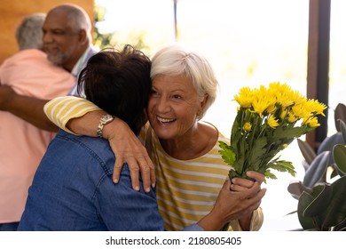 Biracial Happy Senior Guests With Bouquet Embracing Friends At Entrance In Nursing Home. Welcoming, Flower, Greeting, Unaltered, Togetherness, Support, Assisted Living And Retirement Concept.