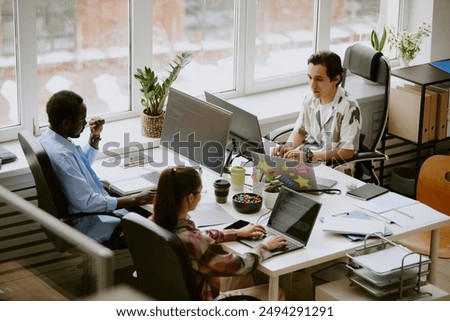 Biracial highly skilled programmer team writing codes surrounded by digital devices, coffee and snacks while sitting at white wooden table in roomy light modern office