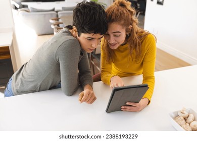 Biracial lesbian couple using tablet, standing leaning on countertop in sunny kitchen. Communication, gay, relationship, togetherness, domestic life and lifestyle, unaltered. - Powered by Shutterstock