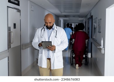 Biracial male doctor wearing lab coat using tablet in corridor at hospital. Hospital, communication, medicine, healthcare and work, unaltered. - Powered by Shutterstock