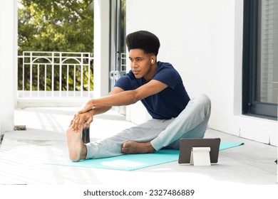 Biracial man doing yoga, stretching and using tablet sitting on sunny balcony. Wellbeing, fitness, communication and healthy lifestyle, unaltered. - Powered by Shutterstock