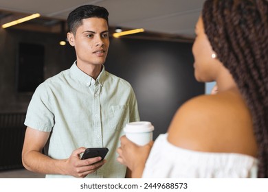 Biracial man holding smartphone talking to biracial woman in a modern business office. Both have dark hair, he in light shirt, she in white, looking relaxed, unaltered. - Powered by Shutterstock