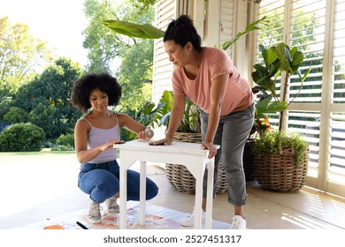 A biracial mother and adult daughter paint furniture outside, both with short hair. Mother hair is black, daughter curly brown, dressed casually, enjoying sunny day, unaltered - Powered by Shutterstock