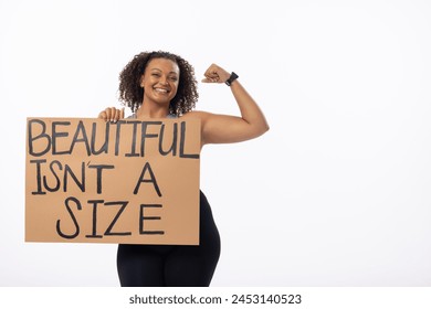 Biracial plus-size young woman flexes arm with a poster on white background, copy space. She sports curly brown hair, a bright smile, and black activewear, promoting body positivity, unaltered - Powered by Shutterstock