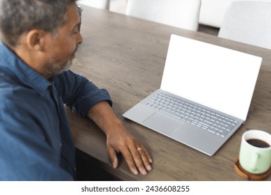 Biracial senior man with graying hair works on a laptop at a wooden table, with copy space. A cup of coffee sits nearby, suggesting a casual work setting at home or a café. - Powered by Shutterstock