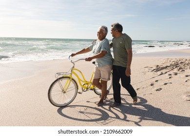 Biracial senior man walking and talking with woman wheeling bicycle at sunny beach against sky. active lifestyle and transportation. - Powered by Shutterstock