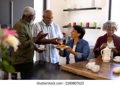 Biracial Smiling Senior Woman Offering Cookies To Multiracial Friends At Table In Nursing Home. Coffee, Snacks, Food, Unaltered, Friendship, Togetherness, Support, Assisted Living And Retirement.