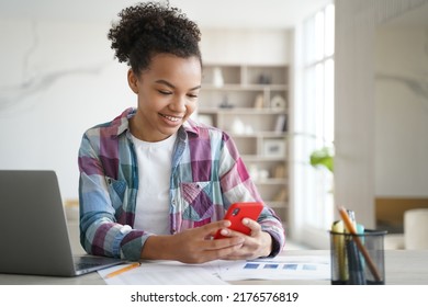 Biracial Teen Girl Chatting Online On Smartphone While Doing Homework. Schoolgirl Holding Phone, Distracted From Studying Learning. New Generations Addicted With Social Networks, Overuse Of Gadgets.