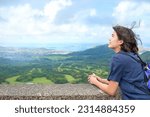 Biracial teen girl standing at top of Pali Lookout enjoying view of Oahu valley over Kaneohe city and Hawaiian ocean in background