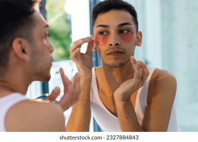 Biracial transgender man looking in mirror and applying under-eye patches in bathroom. Gender, self care, lifestyle and domestic life, unaltered. - Powered by Shutterstock