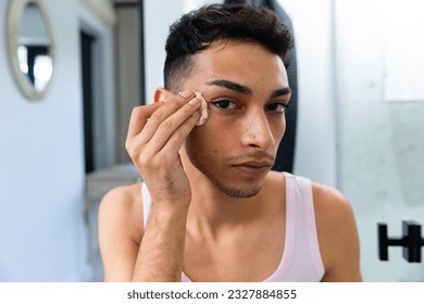 Biracial transgender man looking in mirror, using cotton pad for cleansing face in bathroom. Gender, self care, lifestyle and domestic life, unaltered. - Powered by Shutterstock