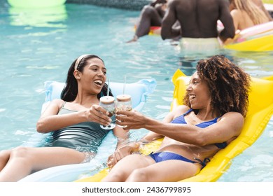 Biracial woman and African American woman lounge on floaties, toasting drinks. They are enjoying a sunny day at a pool party, surrounded by others in the water. - Powered by Shutterstock