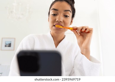 Biracial woman in bathrobe brushing teeth and using smartphone in sunny bathroom. Lifestyle, self care, hygiene, communication and domestic life, unaltered. - Powered by Shutterstock