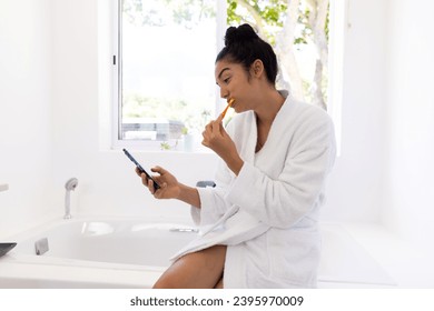 Biracial woman in bathrobe brushing teeth and using smartphone in sunny bathroom. Lifestyle, self care, hygiene, communication and domestic life, unaltered. - Powered by Shutterstock