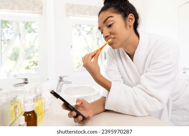 Biracial woman in bathrobe brushing teeth and using smartphone in sunny bathroom. Lifestyle, self care, hygiene, communication and domestic life, unaltered. - Powered by Shutterstock