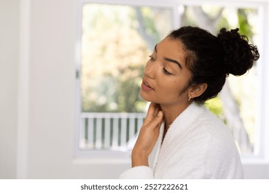 Biracial woman in bathrobe looking in mirror in sunny bathroom. Lifestyle, self care and domestic life, unaltered. - Powered by Shutterstock