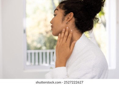 Biracial woman in bathrobe looking in mirror in sunny bathroom. Lifestyle, self care and domestic life, unaltered. - Powered by Shutterstock