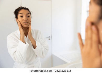 Biracial woman in bathrobe looking in mirror in sunny bathroom. Lifestyle, self care and domestic life, unaltered. - Powered by Shutterstock