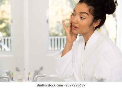 Biracial woman in bathrobe looking in mirror in sunny bathroom. Lifestyle, self care and domestic life, unaltered. - Powered by Shutterstock