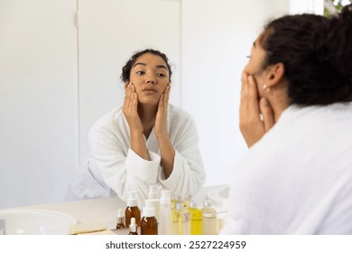 Biracial woman in bathrobe looking in mirror in sunny bathroom. Lifestyle, self care and domestic life, unaltered. - Powered by Shutterstock