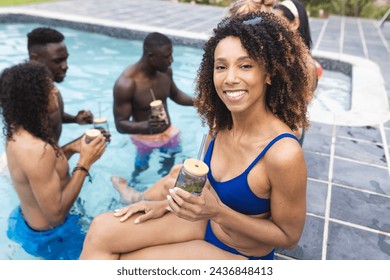 Biracial woman in blue swimwear enjoys a drink by the pool; diverse friends chat in the background. It is a sunny day, perfect for outdoor relaxation and socializing. - Powered by Shutterstock