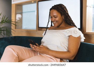 Biracial woman with braided hair using smartphone in a modern business office. She wears a white top, seated comfortably, looking at the screen, unaltered. - Powered by Shutterstock