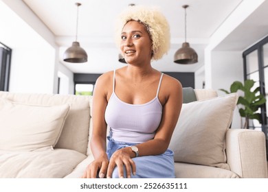 Biracial woman with curly blonde hair in a blue dress and silver watch sits on a sofa. She looks relaxed and thoughtful in a well-lit, modern living room, unaltered - Powered by Shutterstock