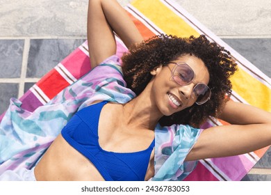 A biracial woman enjoys the sun while lying on a colorful towel. She sports a blue bikini, large sunglasses, and a joyful smile, indicating a leisurely day outdoors. - Powered by Shutterstock