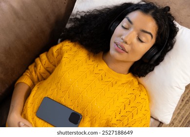 Biracial woman with headphones and smartphone lying on sofa at home. Lifestyle, free time, communication and domestic life, unaltered. - Powered by Shutterstock