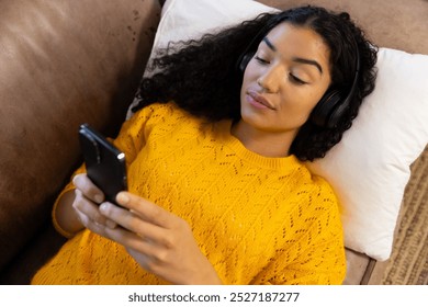 Biracial woman with headphones using smartphone lying on sofa at home. Lifestyle, free time, communication and domestic life, unaltered. - Powered by Shutterstock