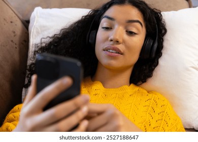 Biracial woman with headphones using smartphone lying on sofa at home. Lifestyle, free time, communication and domestic life, unaltered. - Powered by Shutterstock