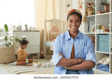 Biracial woman smiling at camera with arms crossed in modern nursery setting Child playing with toys in background and shelves filled with children's items - Powered by Shutterstock