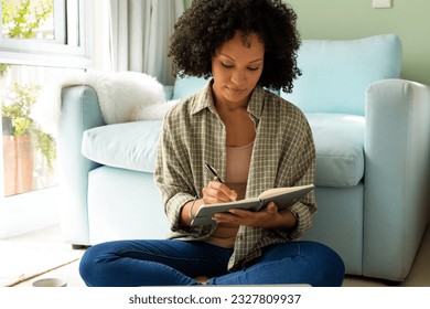 Biracial woman taking notes in notebook sitting on floor in bedroom. Lifestyle and domestic life, unaltered. - Powered by Shutterstock