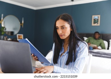 Biracial Woman Using Laptop And Holding Notes, Working In Bedroom, With Partner In Background. Communication, Working From Home, Domestic Life And Inclusivity Concept.