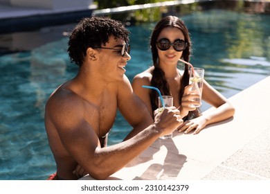 Biracial young couple wearing sunglasses having drinks while enjoying in swimming pool on sunny day. Unaltered, love, togetherness, lifestyle, pool party, summer and weekend activities concept. - Powered by Shutterstock