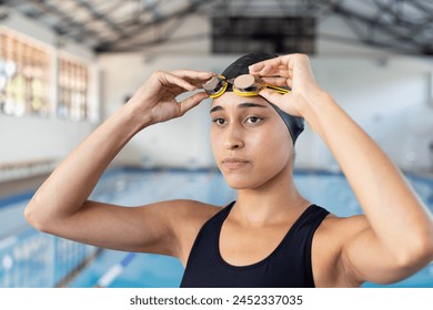 Biracial young female swimmer adjusting goggles at poolside indoors, ready for training. She has light brown skin, dark hair, and is wearing a black swimsuit. - Powered by Shutterstock