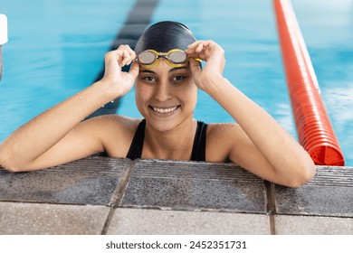 Biracial young female swimmer adjusting goggles at pool edge indoors, smiling. She has light brown skin, dark hair, and brown eyes, wearing a swim cap. - Powered by Shutterstock