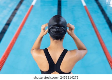 Biracial young female swimmer adjusting swim cap at pool indoors. She has dark hair, wearing a black swimsuit, ready for training, unaltered. - Powered by Shutterstock