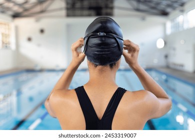 Biracial young female swimmer adjusting swim cap, standing poolside indoors. She has short dark hair, wearing a black swimsuit, ready to dive in, unaltered. - Powered by Shutterstock