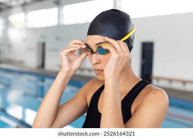 Biracial young female swimmer adjusting goggles indoors, preparing to swim. She has light brown skin, dark hair, and is wearing a black swimsuit, unaltered. - Powered by Shutterstock