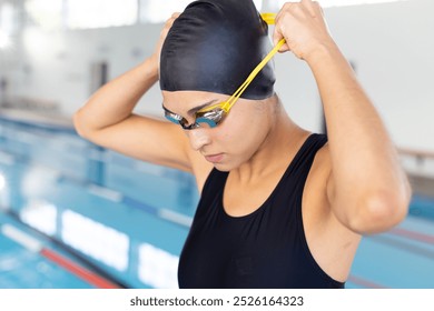 Biracial young female swimmer adjusting goggles indoors, preparing to swim. She has brown skin, dark hair, wearing a black swimsuit, unaltered. - Powered by Shutterstock