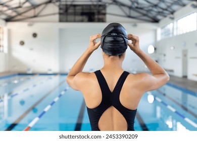 Biracial young female swimmer adjusts her black swim cap at an indoor pool, readying for swim practice. She has dark hair, wearing a black swimsuit, ready to swim, unaltered. - Powered by Shutterstock