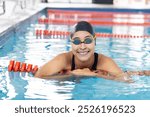 Biracial young female swimmer resting at swimming pool edge, smiling. She, having dark hair and light brown skin, wearing goggles, enjoying calm, unaltered