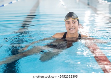 Biracial young female swimmer resting at pool edge indoors, wearing goggles. She has brown hair, light brown skin, and is smiling, unaltered. - Powered by Shutterstock