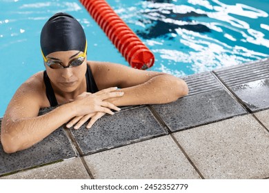 A biracial young female swimmer resting at poolside indoors, wearing goggles. She has dark hair, light brown skin, and is wearing a black swim cap. - Powered by Shutterstock