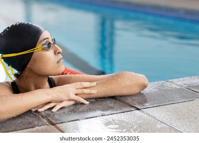 Biracial young female swimmer resting at pool edge indoors, looking thoughtful, copy space. She has light brown skin, dark hair, and is wearing goggles and swim cap, unaltered - Powered by Shutterstock