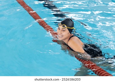 Biracial young female swimmer resting at swimming pool edge indoors, wearing goggles, copy space. She has short brown hair, smiling, looking fit and young, unaltered - Powered by Shutterstock