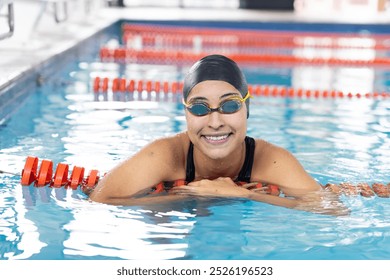 Biracial young female swimmer resting at swimming pool edge, smiling. She, having dark hair and light brown skin, wearing goggles, enjoying calm, unaltered - Powered by Shutterstock